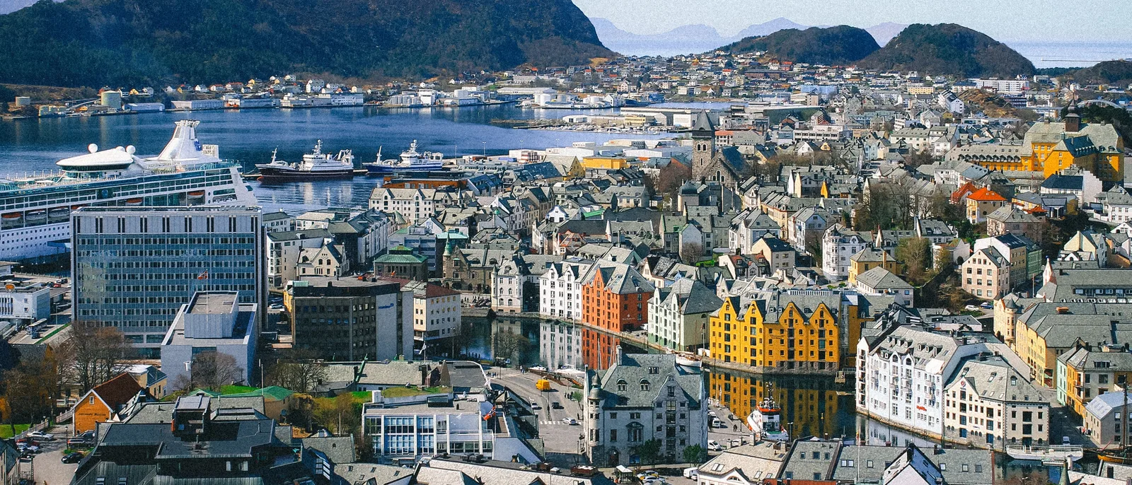 Central Ålesund from above, Art Nouveau buildings in yellow, orange and white lining the Brosundet canal, with a cruise ship in the harbour and mountains across the water.