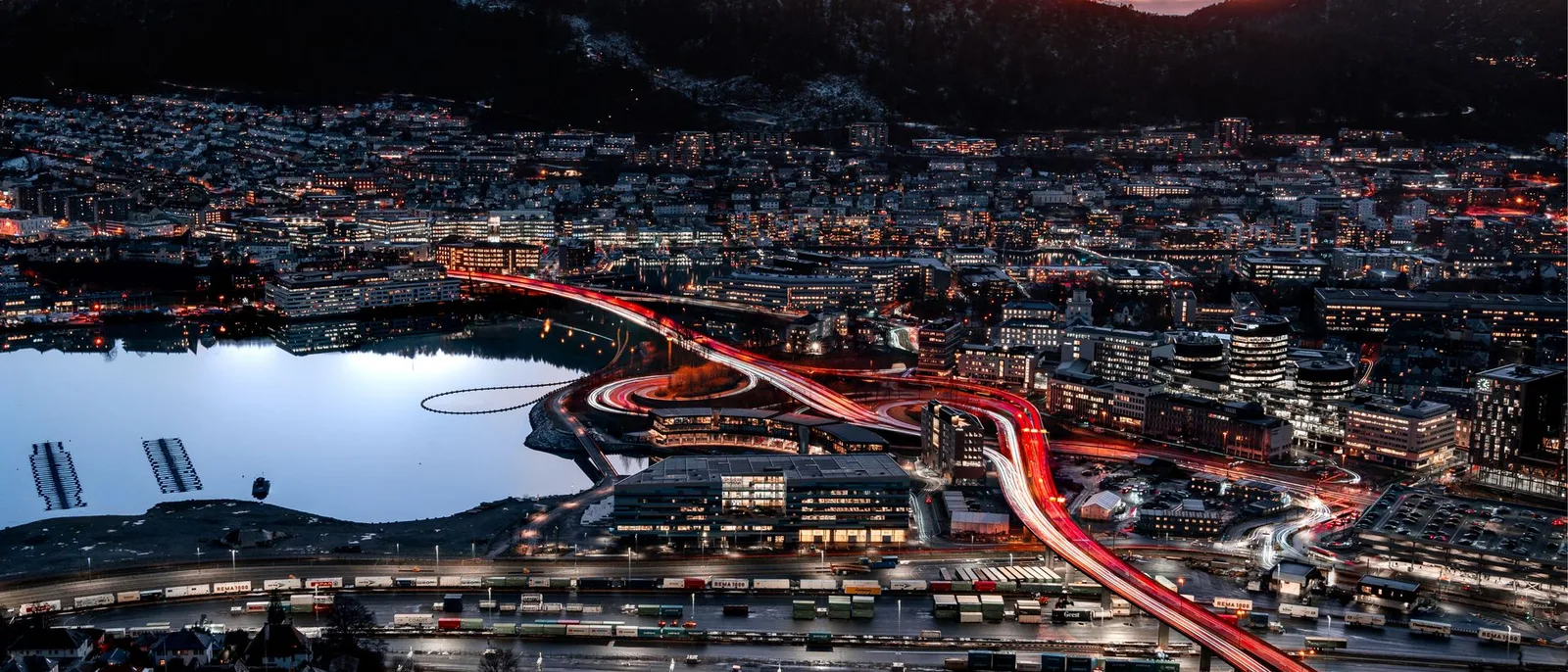 Bergen at dusk from Mount Fløyen, headlight trails on the ring road, the container quay lit below and a pink sky over the hills.