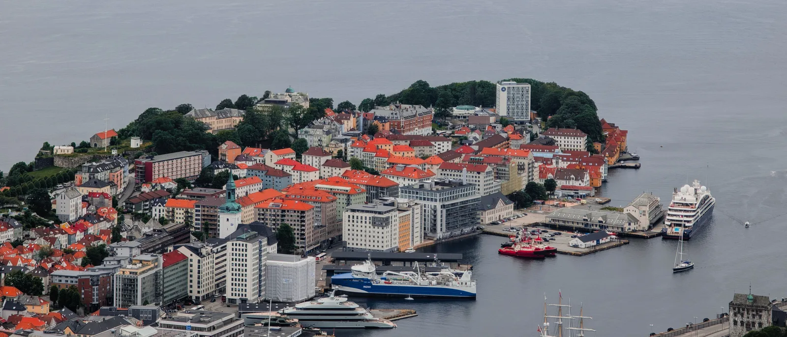 Bergen seen from the air, the Vågen inlet and the peninsula of red-roofed old town buildings, a cruise ship berthed on the far quay and the fjord opening out behind.