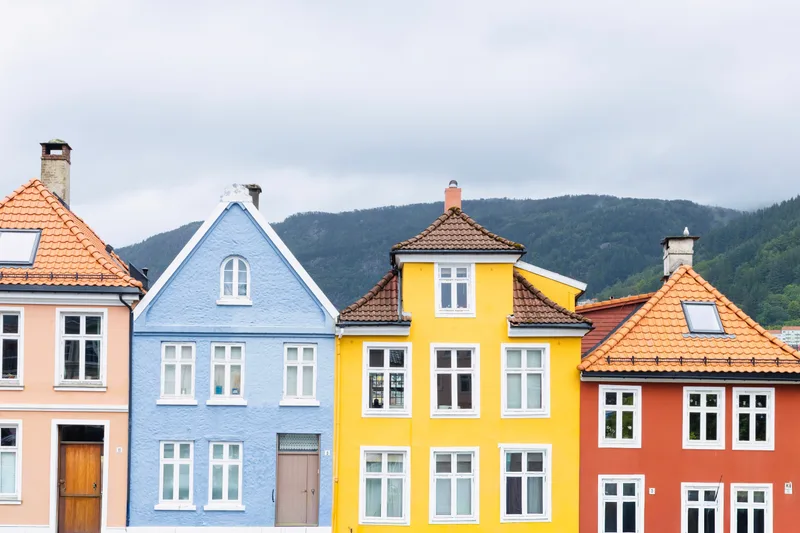 Four painted timber house gables in a row in Bergen, each a different colour and roof shape, with a forested ridge behind them.