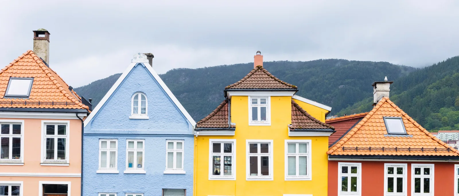 Four painted timber house gables in a row in Bergen, one peach, one blue, one yellow, one red, each with a different roof, a forested ridge rising behind them under a grey sky.