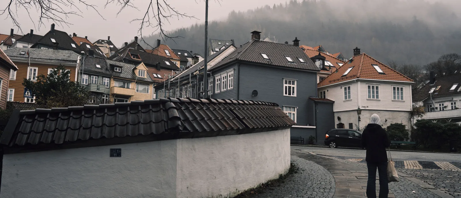 A residential street in Bergen under mist, wooden houses in grey, ochre and white stepping up the hillside behind a whitewashed wall carrying a house number, a woman walking away along wet cobbles.