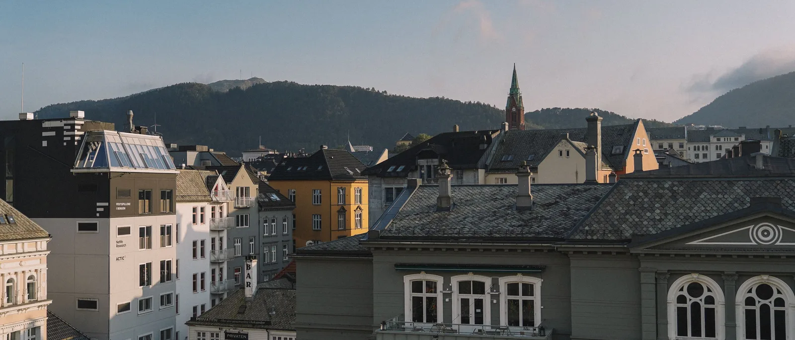 Bergen rooftops in low evening light, slate tiles and a yellow painted townhouse in the foreground, a green copper church spire and a wooded ridge behind.