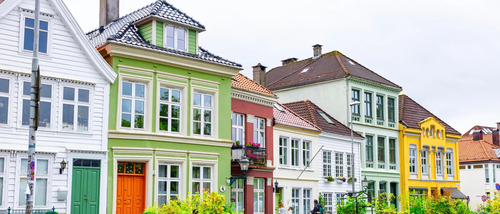 A row of painted wooden houses on a street in Bergen, with white, green, red and yellow facades, tiled roofs and a strip of garden in front, under an overcast sky.