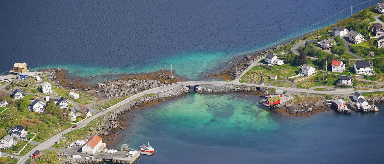 An aerial view of a Lofoten fishing village where a low stone causeway and a small bridge carry a single road between two islands across pale green shallow water, red and white houses scattered on both shores.