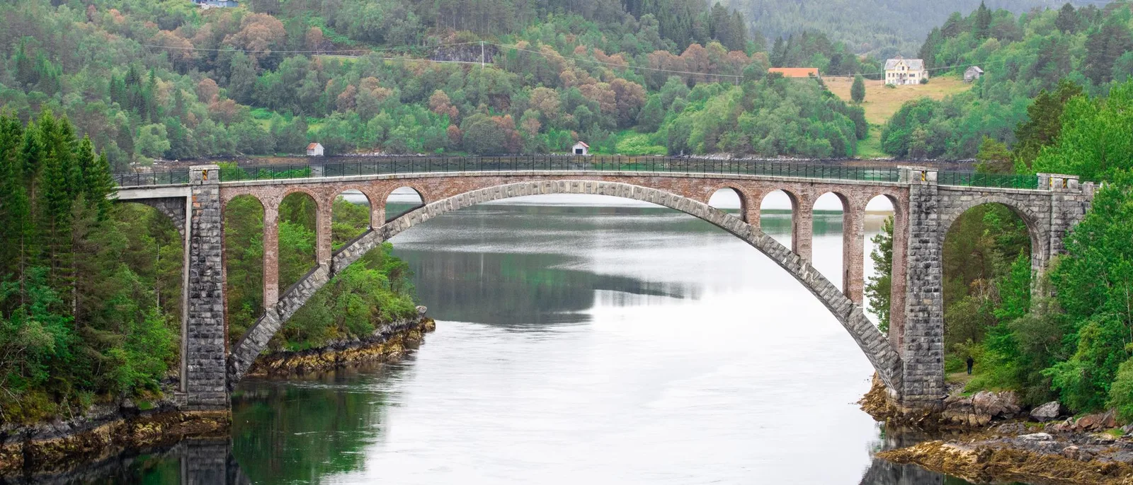 An old stone arch bridge crossing a narrow, still arm of a fjord, wooded hills on both banks and the arch mirrored in the water.