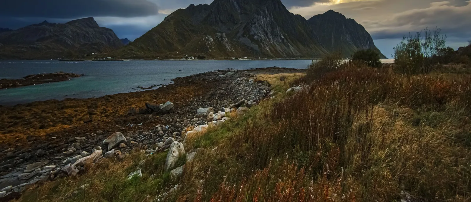 Storm light breaking over a Lofoten shore, a dark peak across the water and a rocky, grassed foreland in the foreground.