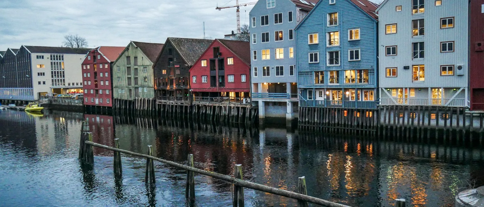 De farvede pakhuse langs Nidelva i Trondheim i skumringen, oplyste vinduer og et trærækværk i forgrunden.